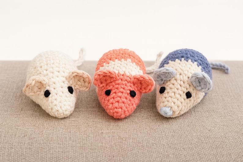 May include: Three handmade crochet mice in a row. One is off-white, one is coral with white stripes, and the last is blue and beige. Each mouse has black eyes, beige ears, and a tail.