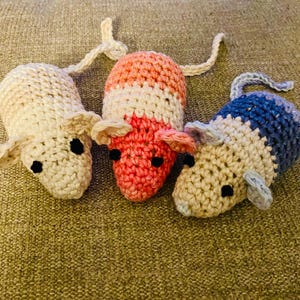 May include: Three handmade crochet mice in cream, pink, and blue. Each mouse has black eyes, pink ears, and a long tail. The mice are made of yarn and are sitting on a textured, brown surface.