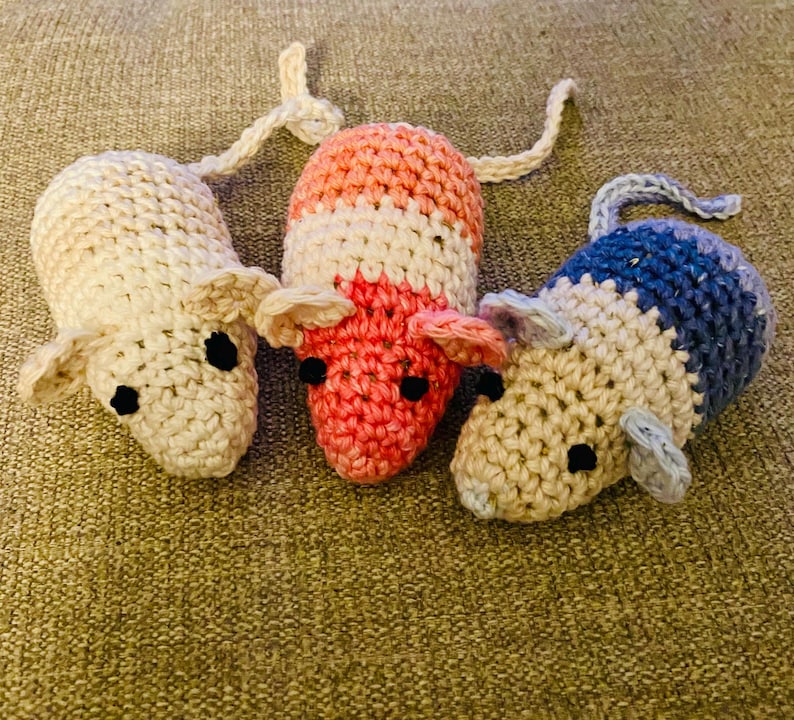 May include: Three handmade crochet mice in cream, pink, and blue. Each mouse has black eyes, pink ears, and a long tail. The mice are made of yarn and are sitting on a textured, brown surface.