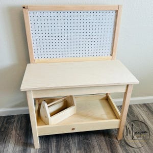 May include: A light-colored wooden desk with a pegboard back and a lower shelf. A small wooden toolbox sits on the shelf. The desk has a natural wood finish and is designed for children's activities.
