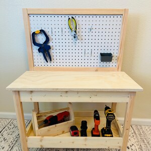 May include: A wooden toy workbench with a pegboard back and a lower shelf. The pegboard holds tools like pliers and a clamp. The shelf contains a wooden toolbox with a red drill and other power tools.