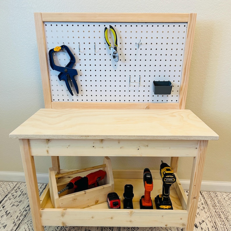 May include: A wooden toy workbench with a pegboard back and a lower shelf. The pegboard holds tools like pliers and a clamp. The shelf contains a wooden toolbox with a red drill and other power tools.