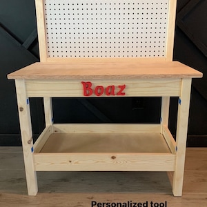May include: A wooden workbench with a pegboard back and a lower shelf. The name "Boaz" is spelled out in red letters on the front. The workbench is made of light-colored wood.