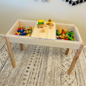May include: A wooden play table with two clear plastic bins filled with colorful wooden blocks. The table has four legs and is sitting on a patterned rug.