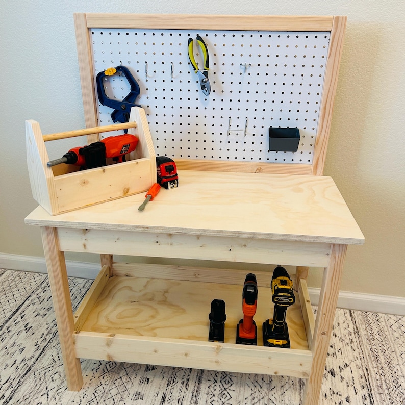 May include: A wooden toy workbench with a pegboard back, a toolbox, and various toy tools. The workbench has a natural wood finish and includes a lower shelf for storage. The tools include a drill, screwdriver, and pliers.