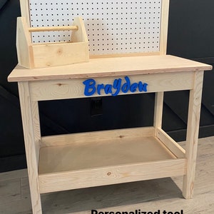 May include: A wooden toy workbench with a pegboard back and a small toolbox. The workbench has a shelf and is personalized with the name "Brayden" in blue. The text at the bottom says "Personalized tool bench with your child's name!"