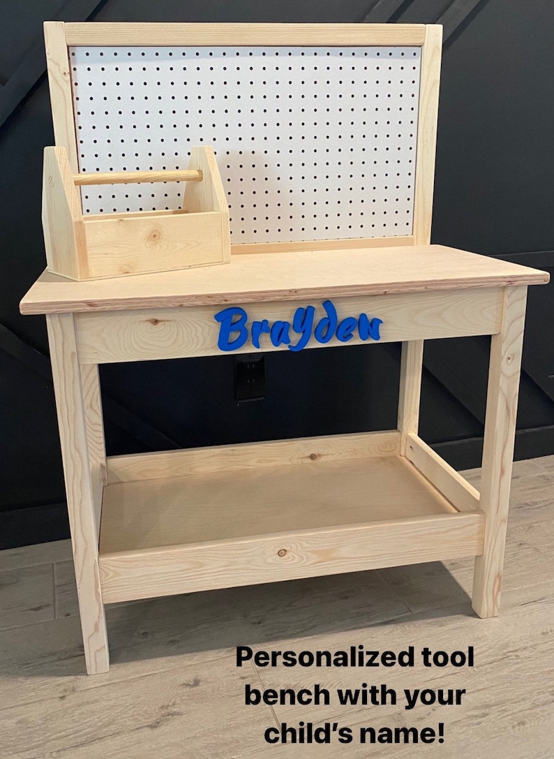 May include: A wooden toy workbench with a pegboard back and a small toolbox. The workbench has a shelf and is personalized with the name "Brayden" in blue. The text at the bottom says "Personalized tool bench with your child's name!"