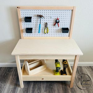 May include: A wooden children's workbench with a pegboard back and a lower shelf. The pegboard holds toy tools, including a hammer, pliers, and shears. Two black storage bins are attached to the pegboard. A wooden toolbox and two toy drills are on the lower shelf.