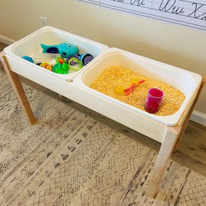 May include: A sensory table with two white rectangular bins supported by light-colored wooden legs. One bin contains a blue whale toy and various colorful plastic shapes. The other bin is filled with yellow corn kernels, a red scoop, and a pink cup.