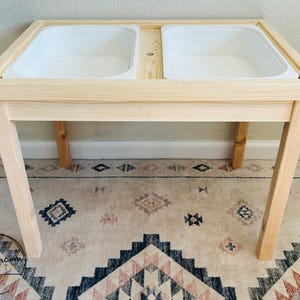 May include: A light-colored wooden sensory table with two white plastic bins. The table has a rectangular frame and angled legs. The table is approximately 30 inches long and 15 inches wide.
