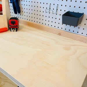 May include: A light-colored wooden workbench with a pegboard back. A red and black tape measure sits on the surface, along with blue pliers. A gray plastic bin and metal hooks are attached to the pegboard.