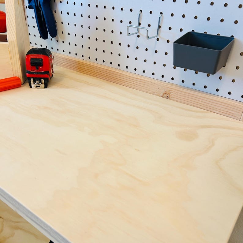 May include: A light-colored wooden workbench with a pegboard back. A red and black tape measure sits on the surface, along with blue pliers. A gray plastic bin and metal hooks are attached to the pegboard.