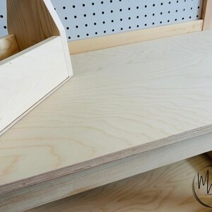 May include: A light-colored wooden desk with a built-in organizer and a drawer. The desk has a natural wood grain finish and a pegboard back. The organizer has multiple compartments for storing items. The drawer has a circular logo.