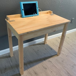 May include: A simple wooden desk with a light wood finish. The desk has a single drawer and a small shelf. The desk is sitting on a light wood floor.