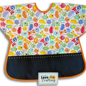 May include: A white bib with a colorful monster print and a black pocket. The bib has orange trim and a yellow stitched line around the pocket. The text "For the Love of Crafting" is on a white tag attached to the bib.