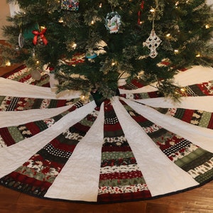 May include: A white quilted Christmas tree skirt with a black trim. The skirt has a patchwork design with red, green, and black fabric. The skirt is decorated with a variety of Christmas patterns.