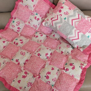 Puede incluir: Una colcha de retazos rosa y blanca con un borde rizado y una almohada rosa y gris con un patrón de chevron. La colcha está hecha de cuadrados con diferentes patrones rosa y blanco, incluyendo lunares, corazones y flamencos.