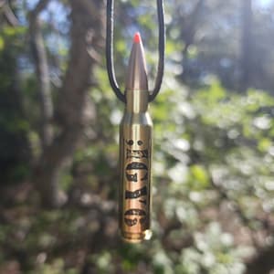 May include: A gold-colored bullet casing necklace with the word "FORGIVE" engraved on it. The necklace is hanging from a black cord.