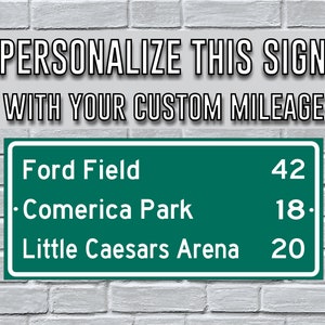 May include: A green road sign with white text that reads "PERSONALIZE THIS SIGN WITH YOUR CUSTOM MILEAGE". Below the text are three lines of text: "Ford Field 42", "Comerica Park 18", and "Little Caesars Arena 20".