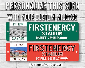 Puede incluir: Dos letreros personalizados con una imagen de estadio, texto que dice "EST. 1999", "CAP. 67,895", "FIRSTENERGY STADIUM", "DISTANCE: 289 MILES", y la información de copyright "signsofwanderlust". Todos los diseños están protegidos por derechos de autor, patente y marca comercial en trámite.