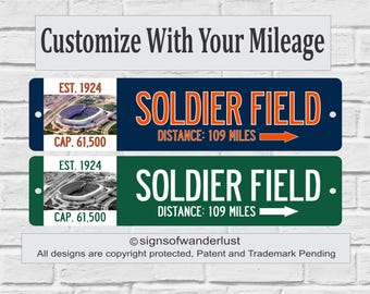 Puede incluir: Dos letreros metálicos rectangulares con el Soldier Field. El letrero superior es azul marino con texto y flecha naranja. El inferior es verde con texto y flecha blancos. Ambos muestran Est. 1924, Cap. 61,500 y Distance: 109 Miles.