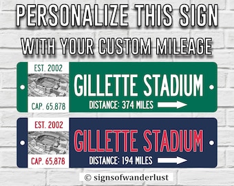 May include: Two personalised metal signs with a green and blue background. The signs feature a photo of Gillette Stadium, the text "EST. 2002", "GILLETTE STADIUM", "CAP. 65,878", and the distance in miles. The signs are customisable with your own mileage.