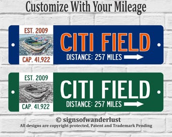 Op de afbeelding: Twee rechthoekige metalen borden met de tekst CITI FIELD, EST. 2009 en CAP. 41.922. Het ene bord is blauw met oranje tekst, het andere groen met witte tekst. Beide bevatten een luchtfoto van het stadion, 257 mijl en een pijl.