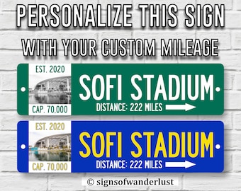 Könnte beinhalten: Zwei personalisierte Metallschildchen mit blauem und grünem Hintergrund. Die Schilder zeigen ein Bild des SoFi Stadiums, den Text "SOFI STADIUM" und den Text "EST. 2020 CAP. 70,000". Die Schilder enthalten auch den Text "DISTANCE: 222 MILES" und einen Pfeil, der nach rechts zeigt.