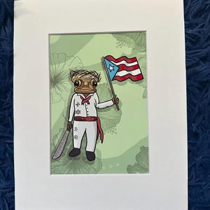 May include: A framed print featuring a cartoon frog in traditional Puerto Rican attire, holding a Puerto Rican flag and two machetes. The frog is dressed in white with a red sash. The background is light green with floral patterns. The text "ESTAMOS AQUI" is on the machetes.