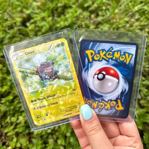 K&ouml;nnte beinhalten: Zwei Pok&eacute;mon-Sammelkarten. Die erste Karte ist gelb und zeigt einen Comic-Frosch mit dem Text "BASIC Coqui 60" und den Attacken "Grass Whistle" und "Echoed Voice". Die zweite Karte ist blau und zeigt das Pok&eacute;mon-Logo und einen Pok&eacute;ball.