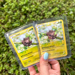 K&ouml;nnte beinhalten: Zwei Pok&eacute;mon-Sammelkarten mit dem Pok&eacute;mon Coqui. Die Karten sind gelb und gr&uuml;n mit einem Bild eines Frosches auf jeder Karte. Die Karten haben den Text "Grass Whistle" und "Echoed Voice" auf sich.