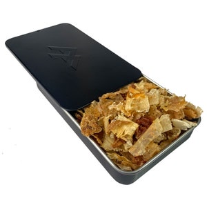 May include: A black metal tin with a logo on the lid is open, revealing a pile of dried, golden-brown wood shavings.