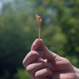 Op de afbeelding: Een brandende lucifer tussen vingers gehouden, met een kleine vlam en rook. De lucifer is van hout met een oranje kop. De achtergrond is een wazig groen, wat duidt op een buitenomgeving.