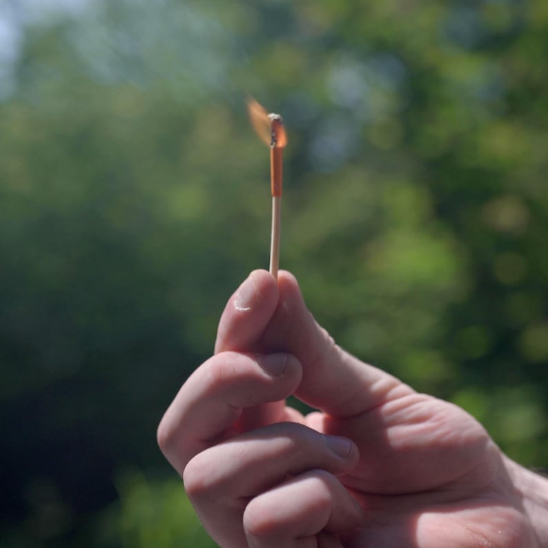Op de afbeelding: Een brandende lucifer tussen vingers gehouden, met een kleine vlam en rook. De lucifer is van hout met een oranje kop. De achtergrond is een wazig groen, wat duidt op een buitenomgeving.