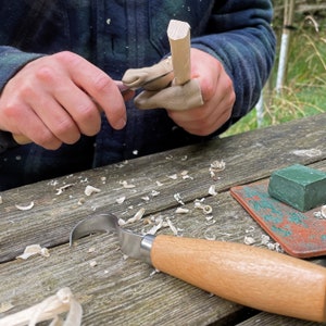 May include: A person is carving a wooden piece with a knife and a carving tool. The wood is light brown and the carving tool has a wooden handle. There are wood shavings on the table.