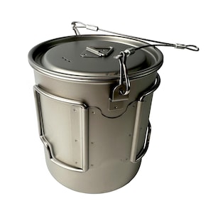 May include: A silver-colored camping pot with a lid and a wire handle. The pot has a cylindrical shape with a curved handle on the side. The lid has a small triangular handle and three small holes. The wire handle is attached to the lid and the side of the pot.