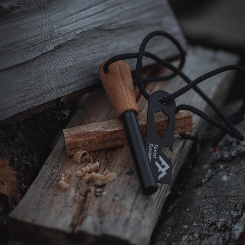 Puede incluir: Un iniciador de fuego negro con un mango de madera y un cord&oacute;n negro. El iniciador de fuego est&aacute; sobre un trozo de madera con virutas de madera. El iniciador de fuego tiene el texto "Forest Fundam" en &eacute;l.