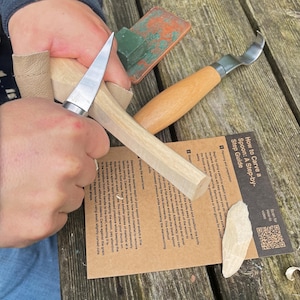 May include: A wood carving setup featuring a carving knife, a curved spoon knife, and a wooden spoon blank. The image shows a step-by-step guide for carving a spoon. The tools are on a wooden surface.