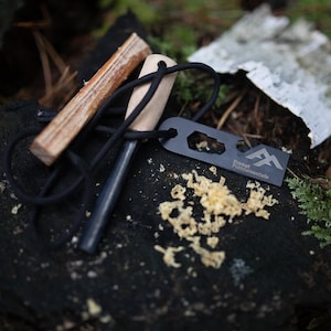 Puede incluir: Un iniciador de fuego negro con un mango de madera y un cord&oacute;n negro. El iniciador de fuego est&aacute; sobre una superficie marr&oacute;n oscura con virutas de madera. El iniciador de fuego tiene el texto "Forest Fundamentals" en &eacute;l.