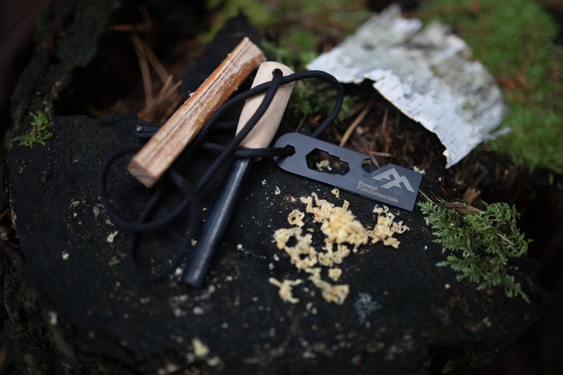 Puede incluir: Un iniciador de fuego negro con un mango de madera y un cord&oacute;n negro. El iniciador de fuego est&aacute; sobre una superficie marr&oacute;n oscura con virutas de madera. El iniciador de fuego tiene el texto "Forest Fundamentals" en &eacute;l.