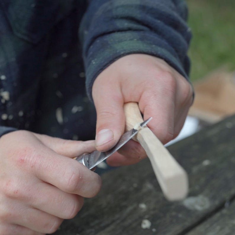 May include: A person carving wood with a silver knife. The wood is light-colored and being shaped with precision. The person is wearing a dark blue and green plaid shirt. The background is blurred, suggesting an outdoor setting.