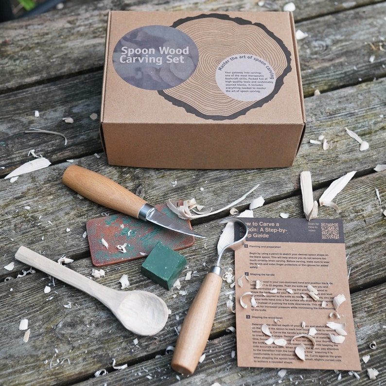May include: A brown cardboard box with the words "Spoon Wood Carving Set" printed on the front. The box is open and contains a set of wood carving tools, including a knife, a gouge, and a sanding block. There is also a piece of wood that has been carved into a spoon shape. The tools are laid out on a wooden table with wood shavings scattered around them. The box also includes a guide on how to carve a spoon.