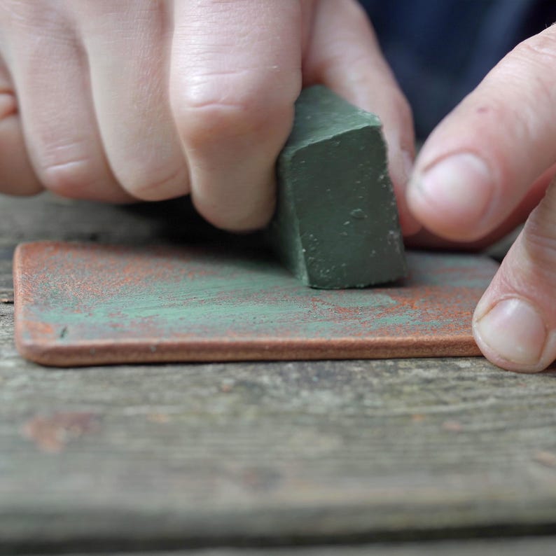 May include: A person's hands are shown using a green block to apply a green pigment to a rectangular, brown surface. The surface has a textured appearance with hints of green. The image is a close-up, focusing on the process of applying the pigment.