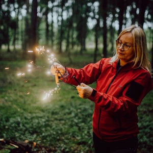 Puede incluir: Una mujer con una chaqueta roja y pantalones negros sostiene un  fuego artificial en un entorno boscoso. El fuego artificial est&aacute; encendido y produce chispas.