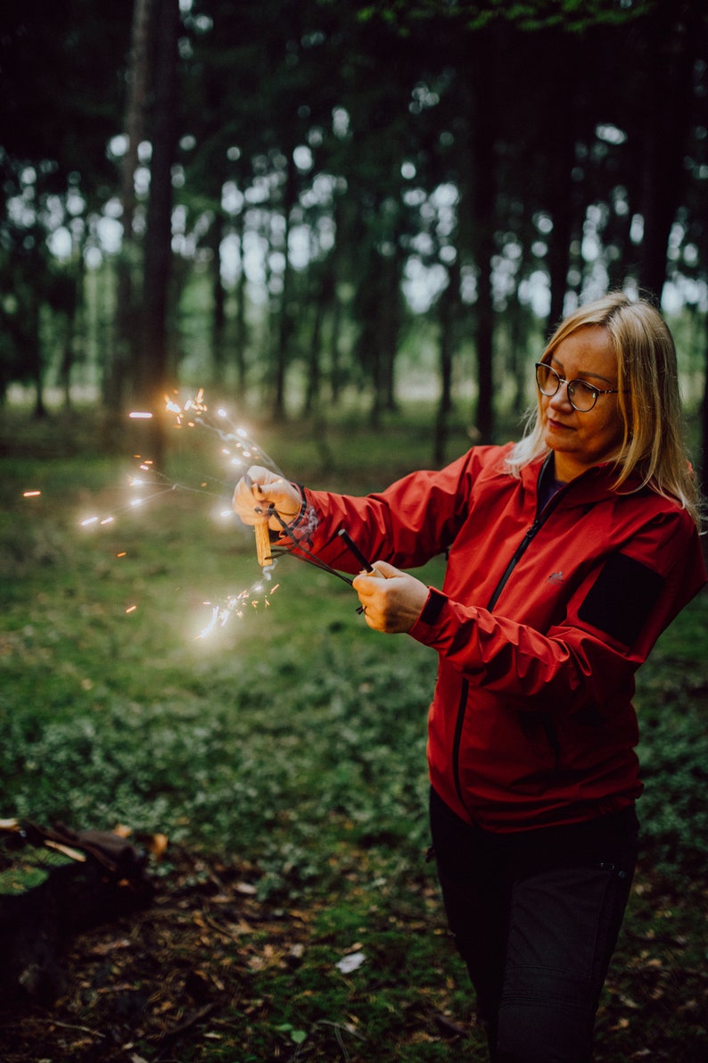 Puede incluir: Una mujer con una chaqueta roja y pantalones negros sostiene un  fuego artificial en un entorno boscoso. El fuego artificial est&aacute; encendido y produce chispas.