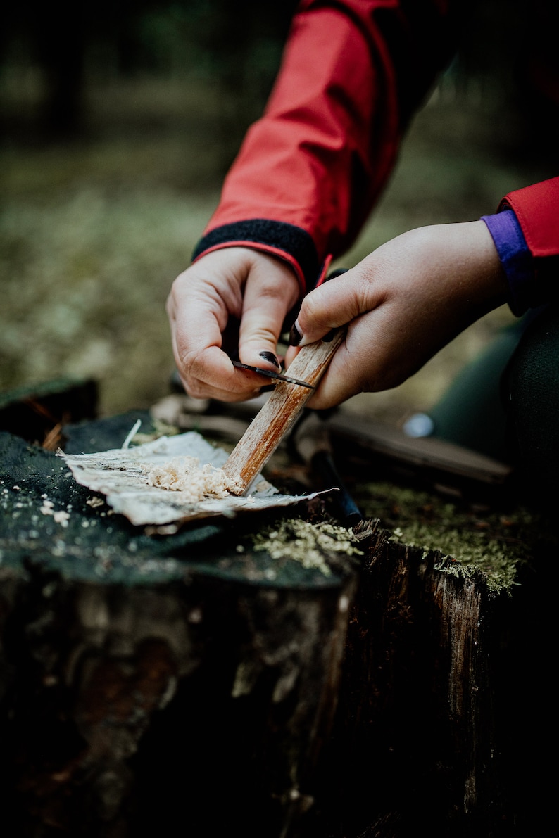 Puede incluir: Una persona utiliza un cuchillo para raer madera en un mont&oacute;n de virutas sobre un toc&oacute;n de &aacute;rbol. La persona lleva una chaqueta roja y sostiene un palo de madera.