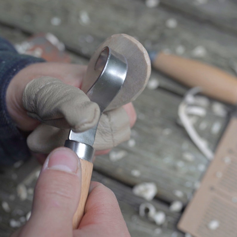 May include: A close-up of a person using a wood carving tool with a wooden handle and a metal blade. The tool is being used to carve a piece of wood.
