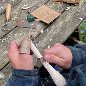 May include: A person is carving a piece of wood with a knife. They are wearing leather finger guards to protect their fingers. The wood is light brown and the knife is silver. There are wood shavings on the table.