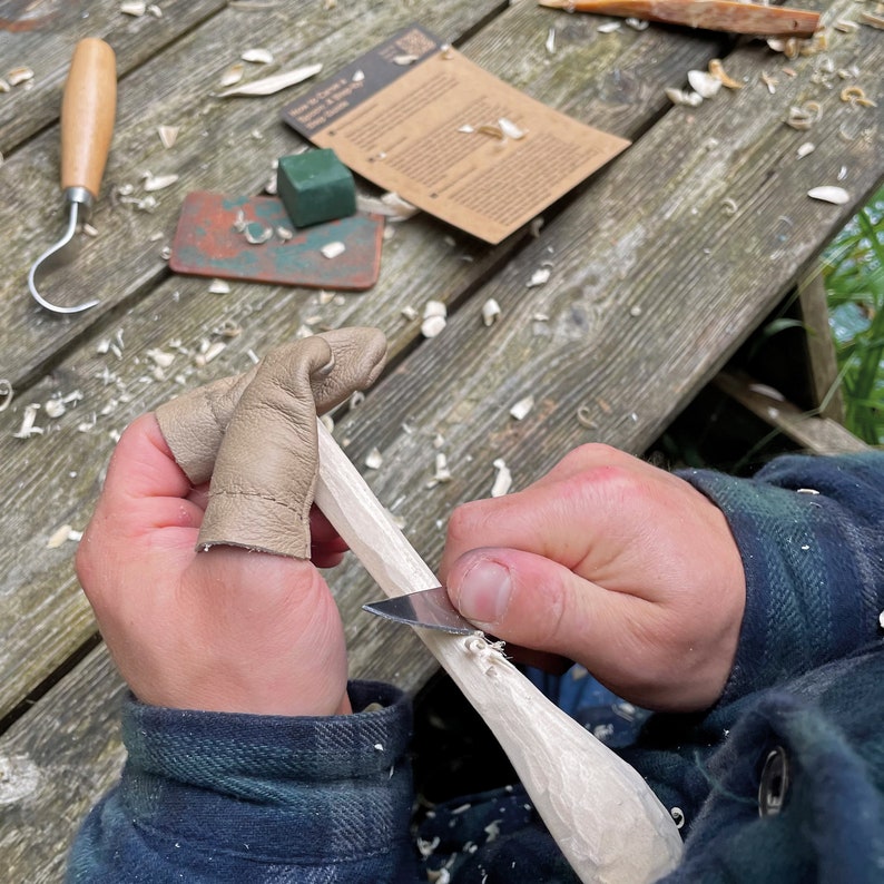 May include: A person is carving a piece of wood with a knife. They are wearing leather finger guards to protect their fingers. The wood is light brown and the knife is silver. There are wood shavings on the table.