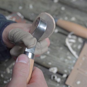 May include: A close-up of a wood carving tool with a curved blade, held in gloved hands. The tool is cutting into a small, round piece of wood. The handle is light-colored wood.